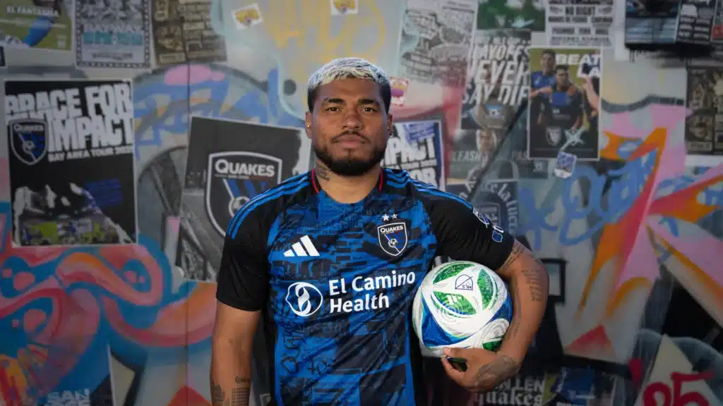 A soccer player in a San Jose Earthquakes jersey holds a ball while standing in front of a graffiti-covered wall with team posters.